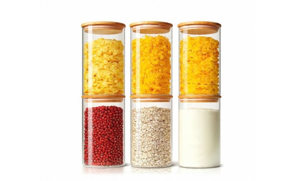 Clear glass storage containers with wooden lids holding cereal, grains, and dry pantry items