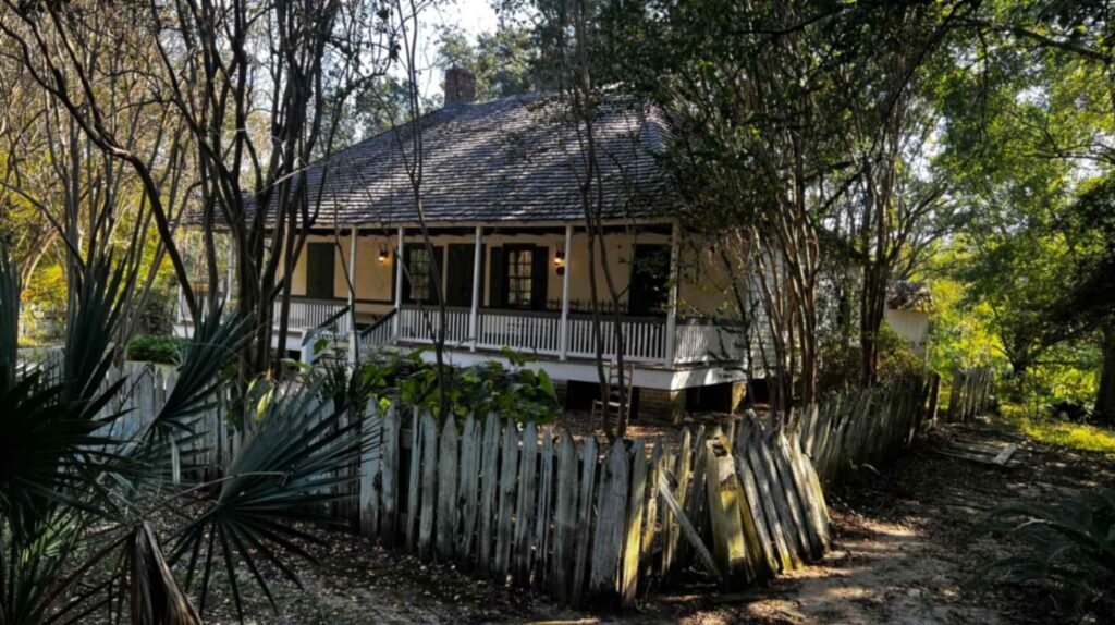 A historic 19th-century Cajun raised cottage with a steep gabled roof, wide front porch, and dark wooden shutters, surrounded by dense trees and a weathered white picket fence.