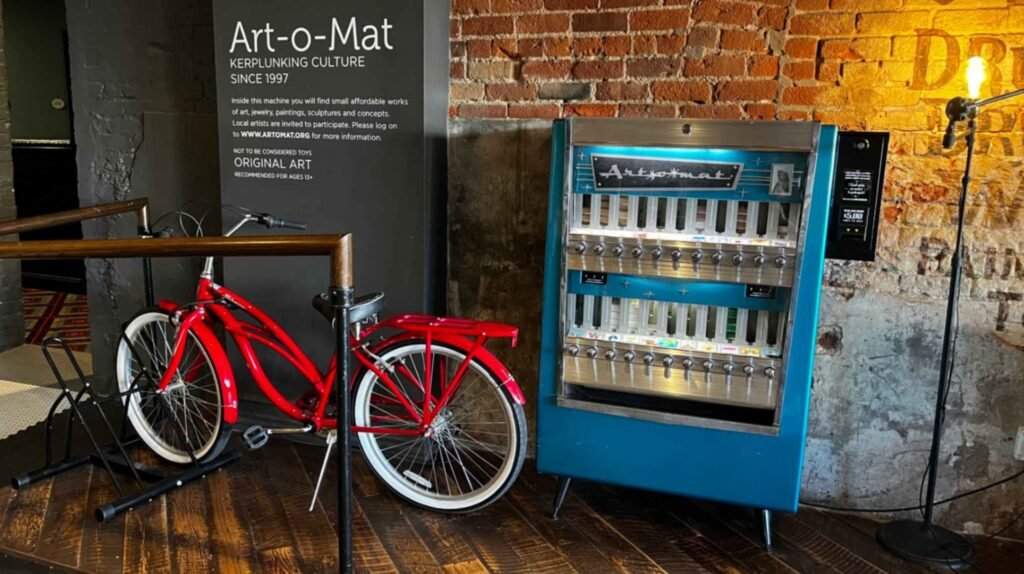 A vintage teal Art-o-Mat vending machine stands next to a red cruiser bicycle against an exposed brick wall in the lobby of The Old No. 77 Hotel & Chandlery.