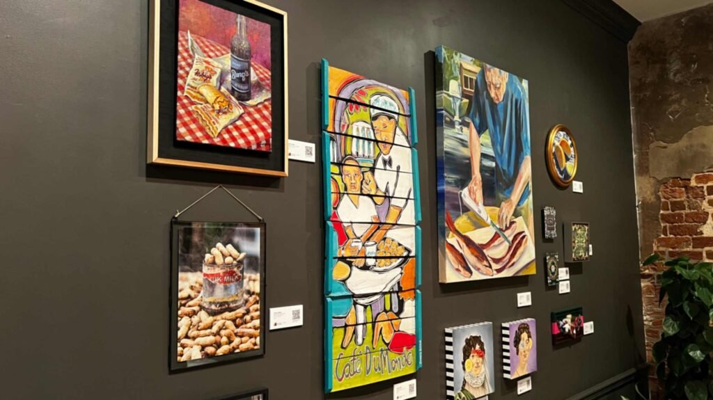 Multiple framed and canvas paintings depicting classic New Orleans food traditions hang grouped together on a dark gray gallery wall inside a building corridor.