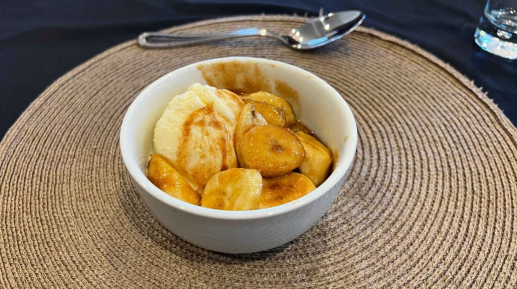 A white bowl filled with freshly prepared Bananas Foster dessert combining sliced bananas, warm caramel sauce, and vanilla ice cream on a round woven placemat.