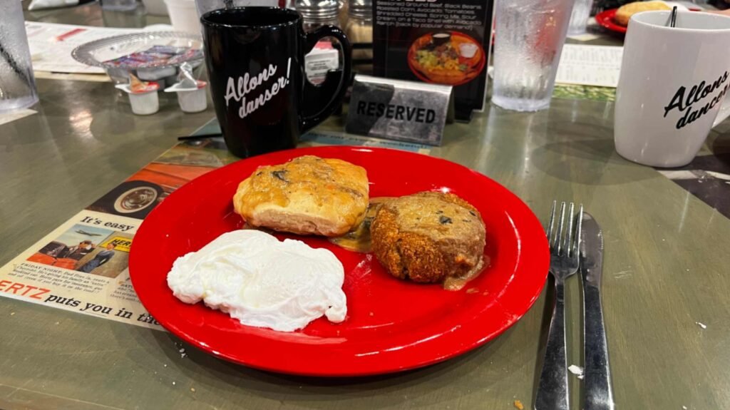 A breakfast plate with a biscuit, boudin ball, gravy, and eggs served next to "Allons danser!" coffee mugs on a restaurant table.