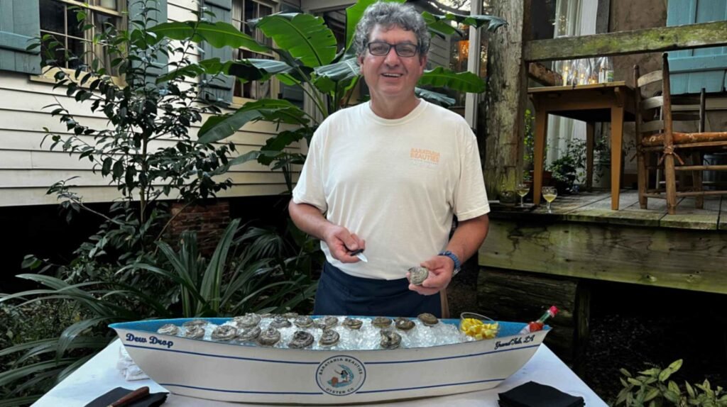 Oyster farmer Albert "Buzzy" Besson smiles while holding a shucked shell above a boat-shaped ice display filled with fresh Barataria Beauties oysters at Maison Madeleine.