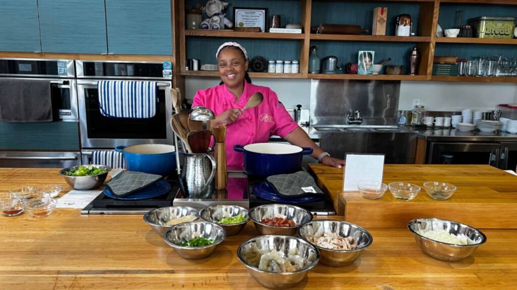 Chef Dee Lavigne smiles behind a wooden cooking station filled with bowls of fresh gumbo ingredients at the Deelightful Roux School of Cooking.
