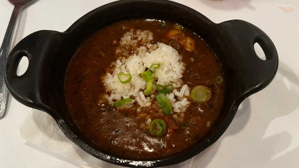 A black cast-iron bowl filled with a dark, smoky Cajun gumbo topped with white rice and sliced green onions.