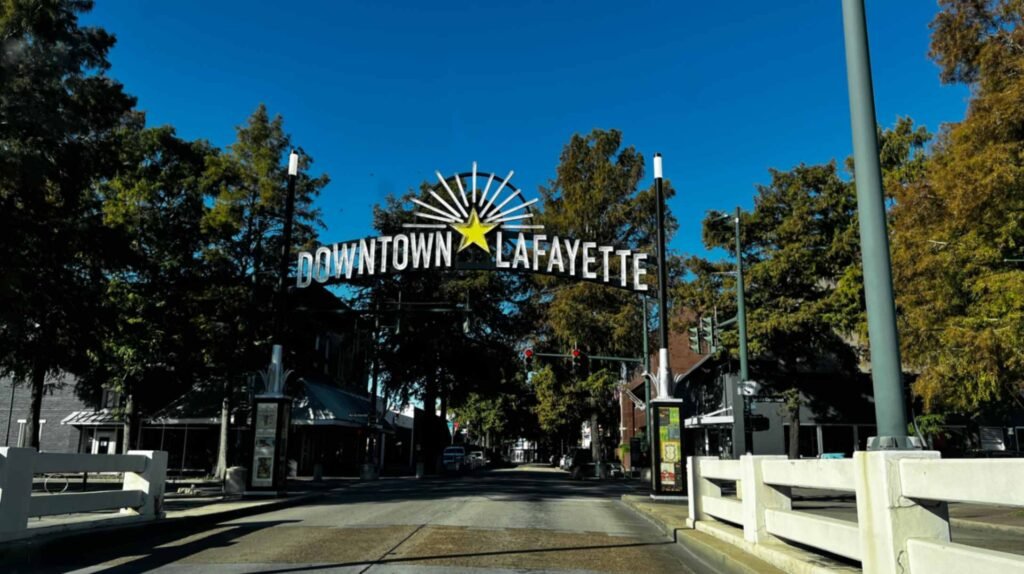 The metal archway sign reading "Downtown Lafayette" spans across a roadway entrance under a clear blue sky, flanked by green trees.