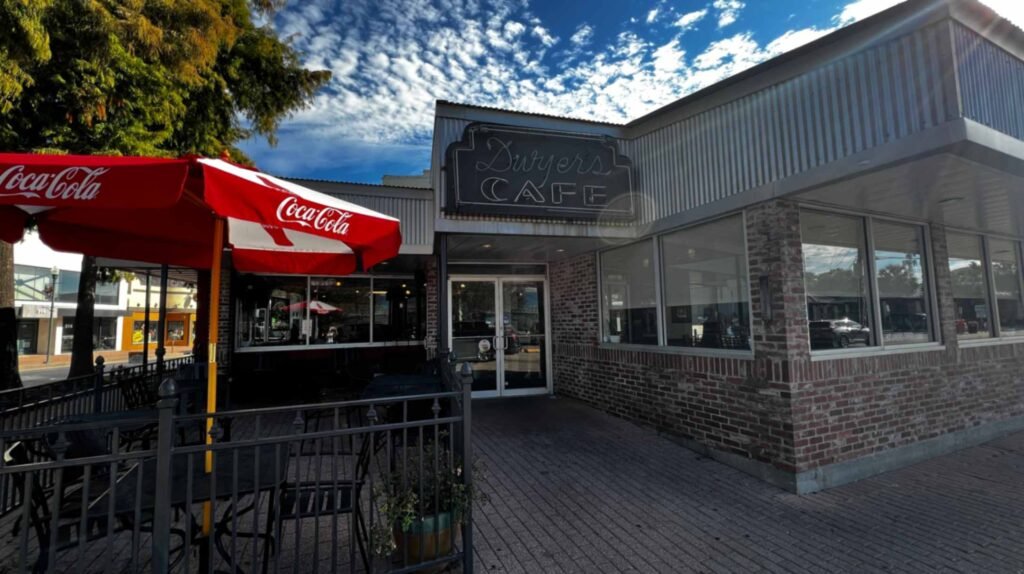 The brick storefront and glass entrance doors of Dwyer's Cafe under an unlit sign, featuring an outdoor patio dining area shaded by red Coca-Cola umbrellas.