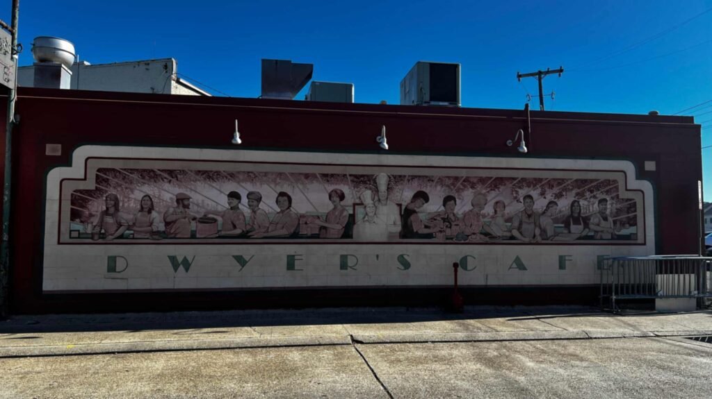 A large outdoor painted mural on the side wall of Dwyer's Cafe depicting a long line of restaurant workers and cooks against a deep red building backdrop.