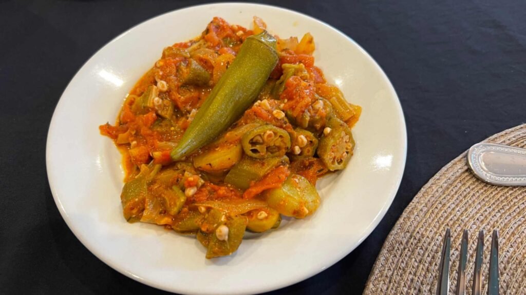 A white plate containing a serving of smothered okra cooked with stewed tomatoes and onions on a black tablecloth.