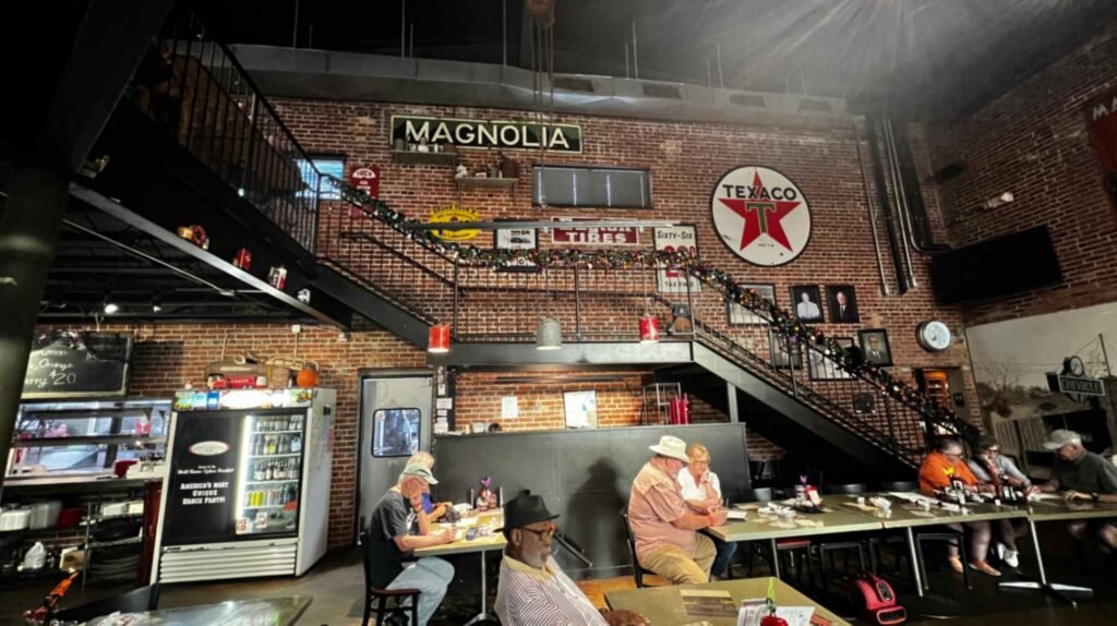 Diners sit at long tables inside the industrial brick interior of Buck & Johnny's restaurant in Breaux Bridge, featuring a metal staircase decorated with vintage Texaco and Magnolia petroleum signs.