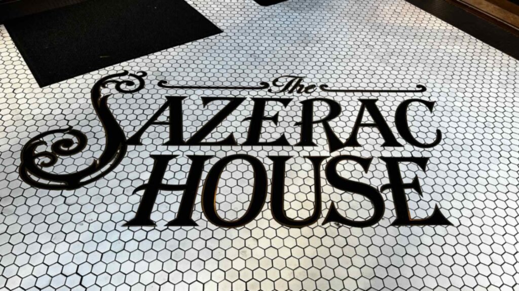 The black script and serif logo reading "The Sazerac House" embedded into a white hexagonal mosaic tile entryway floor.