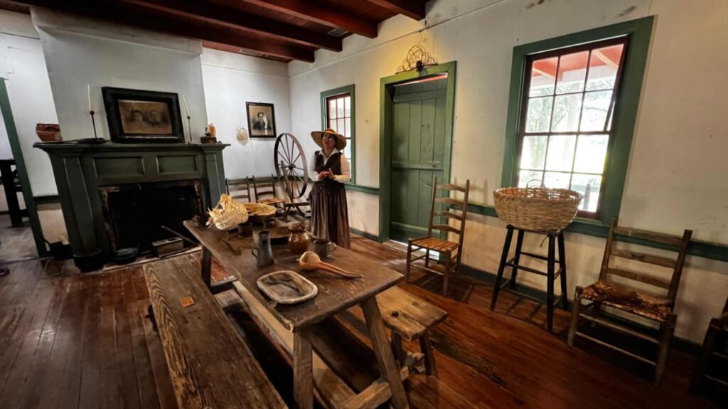 A living history interpreter in period dress stands inside a historic Acadian home featuring a fireplace, a long wooden table with benches, and a spinning wheel at Vermilionville.