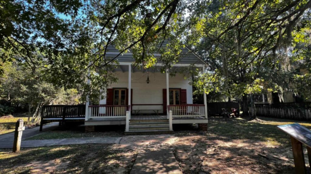 The exterior facade of a historic raised Acadian cottage with red shutters, a wide front porch, and a steep roof shaded by mature live oak trees at Vermilionville.