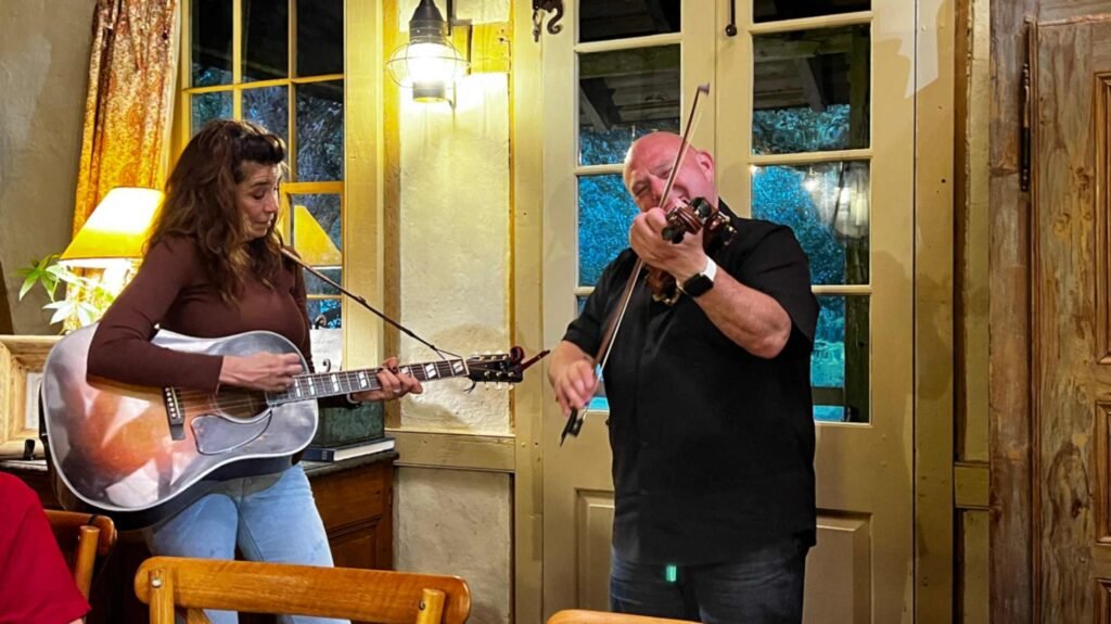 Cajun musicians Yvette Landry playing an acoustic guitar and Beau Thomas playing a fiddle on an indoor porch setting at Maison Madeleine.
