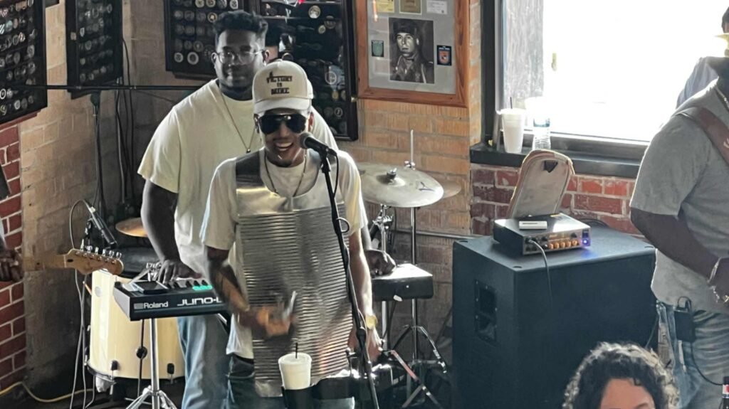 A musician wearing a metal frottoir vest rubboard over his chest sings into a microphone while performing with a Zydeco band at Buck & Johnny's.