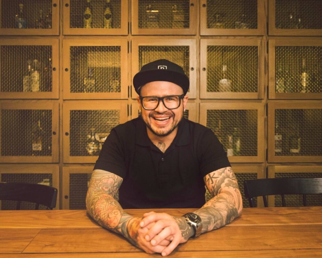 A portrait of a smiling man with arm tattoos wearing a black polo shirt, black glasses, and a black baseball cap. He sits at a wooden table with his hands clasped, positioned in front of a warm wood wall lined with mesh fronted liquor cabinets.