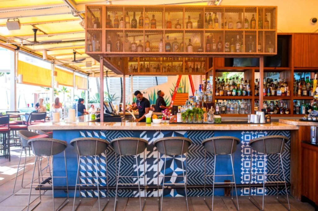 An open air resort bar featuring a blue counter fronted with bold black and white geometric tile patterns. Woven barstools with thin metal legs line the stone countertop, while bartenders prepare drinks beneath an overhead gold mesh liquor display shelf under a yellow awning patio.