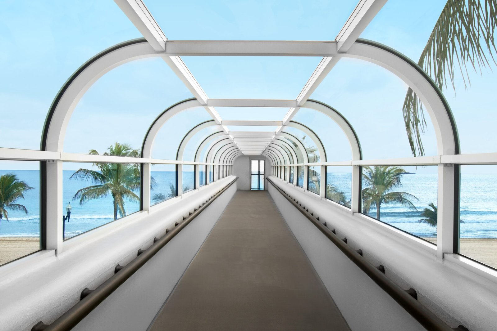 An interior view of a long enclosed glass skywalk bridge with a curved structural white frame. The pedestrian walkway overlooks Fort Lauderdale Beach, offering clear views of palm trees, sandy shores, and blue ocean waves under a bright sky.
