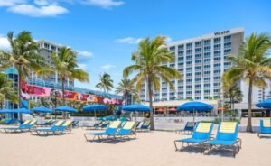 Oceanfront exterior of The Westin Fort Lauderdale Beach Resort & Spa showing the signature skywalk bridge, tropical palm trees, and blue beach chairs on the sand.