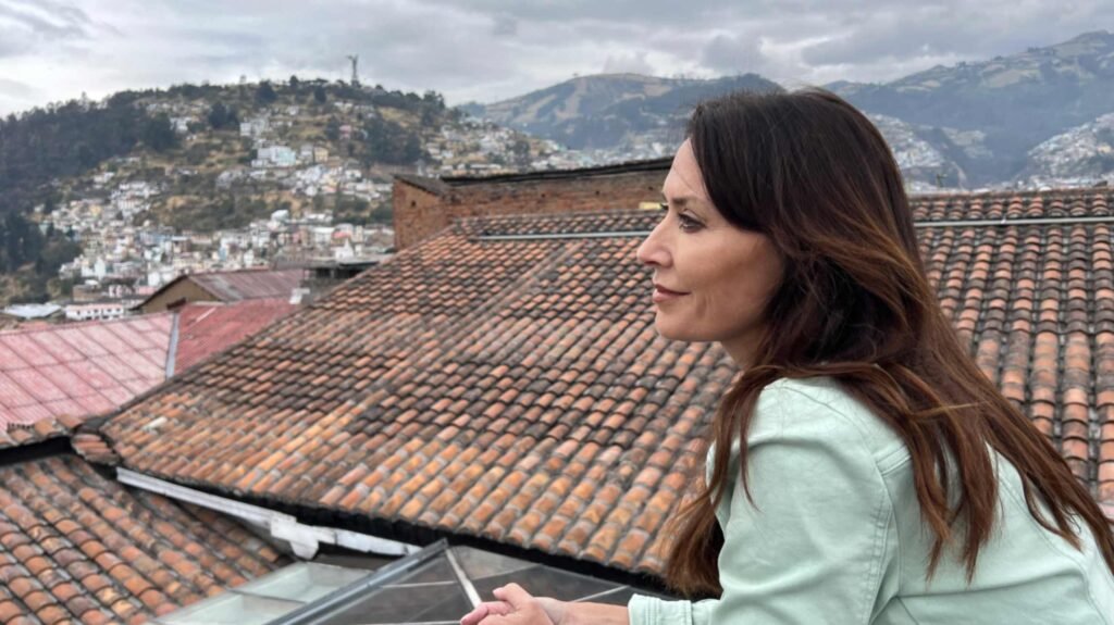 Close up profile view of a woman with long brown hair wearing a light green jacket looking out over rows of weathered orange clay roof tiles in Quito Ecuador with hillsides in the background