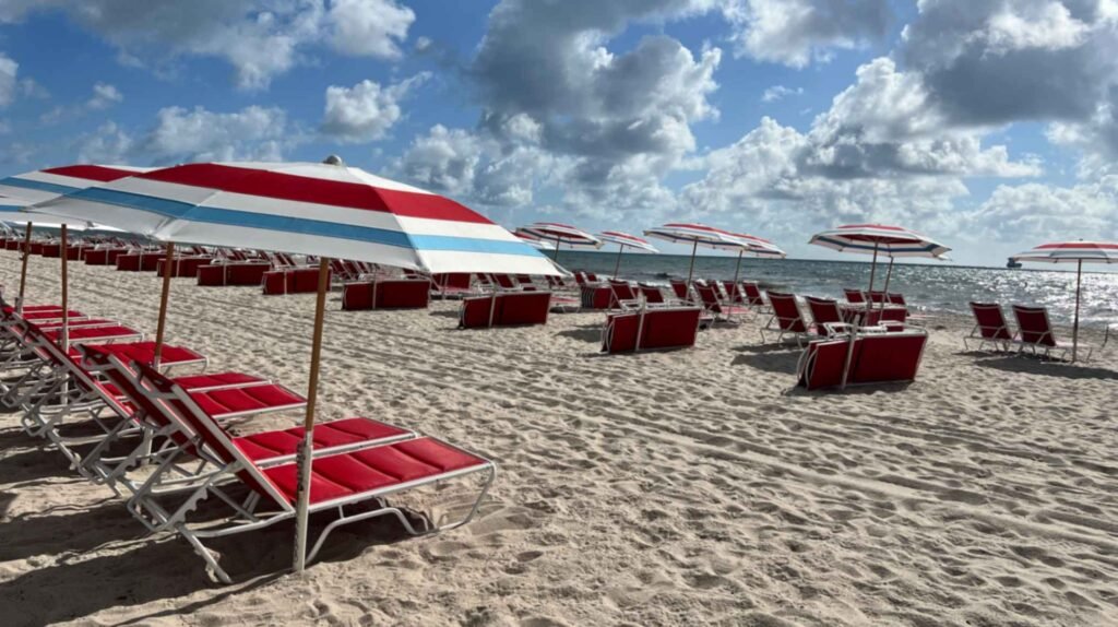 Rows of bright red lounge chairs paired with white, red, and blue striped umbrellas on a wide sandy beach. The beach setup stretches along the Atlantic coastline under a dynamic sky filled with fluffy white and grey clouds. The sun glints off the ocean water in the background.