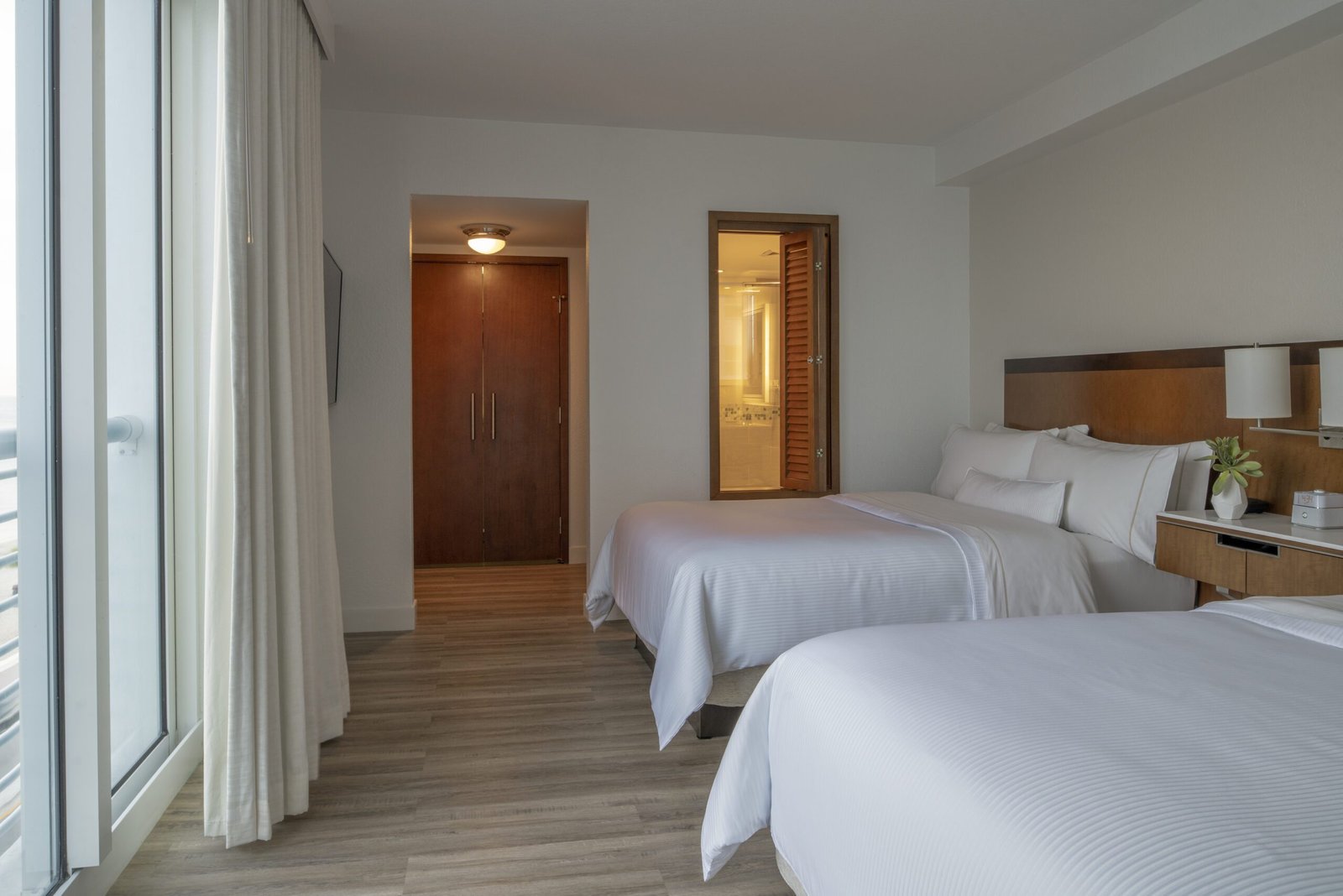 A hotel guest room featuring two queen beds with white linens and a wooden headboard. On the left, floor-to-ceiling glass sliding doors are framed by white curtains. In the background, an open entryway leads to a wooden closet door, and a unique vertical window with wooden shutters looks directly into the illuminated bathroom.