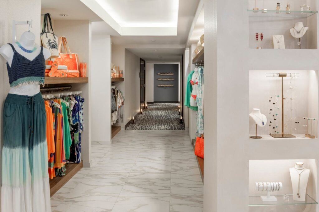 A brightly lit resort boutique corridor with white marble-patterned tile flooring. On the left, clothing racks hold vibrant orange and blue resort wear, alongside a mannequin dressed in a blue knit top and matching ombré skirt. On the right, recessed wall niches with glass shelves showcase delicate jewelry pieces, while the hallway leads down toward a grey wall with modern decor elements.