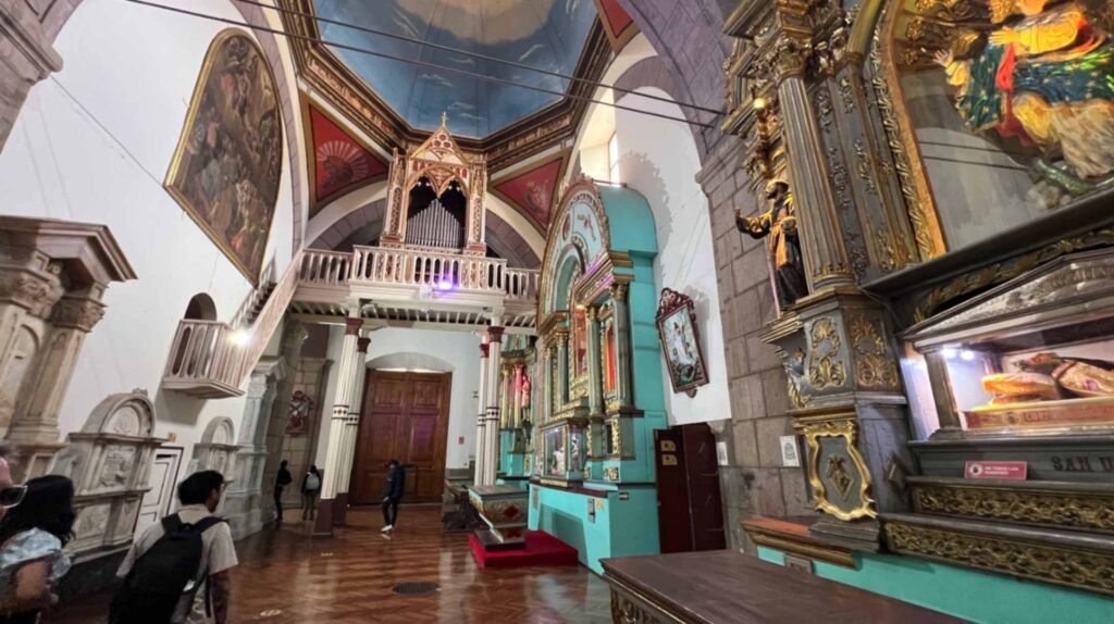Wide interior shot of a grand historic church in Quito looking down a wooden floor corridor toward a raised choir loft with a decorative organ pipe structure under a vaulted ceiling painted blue