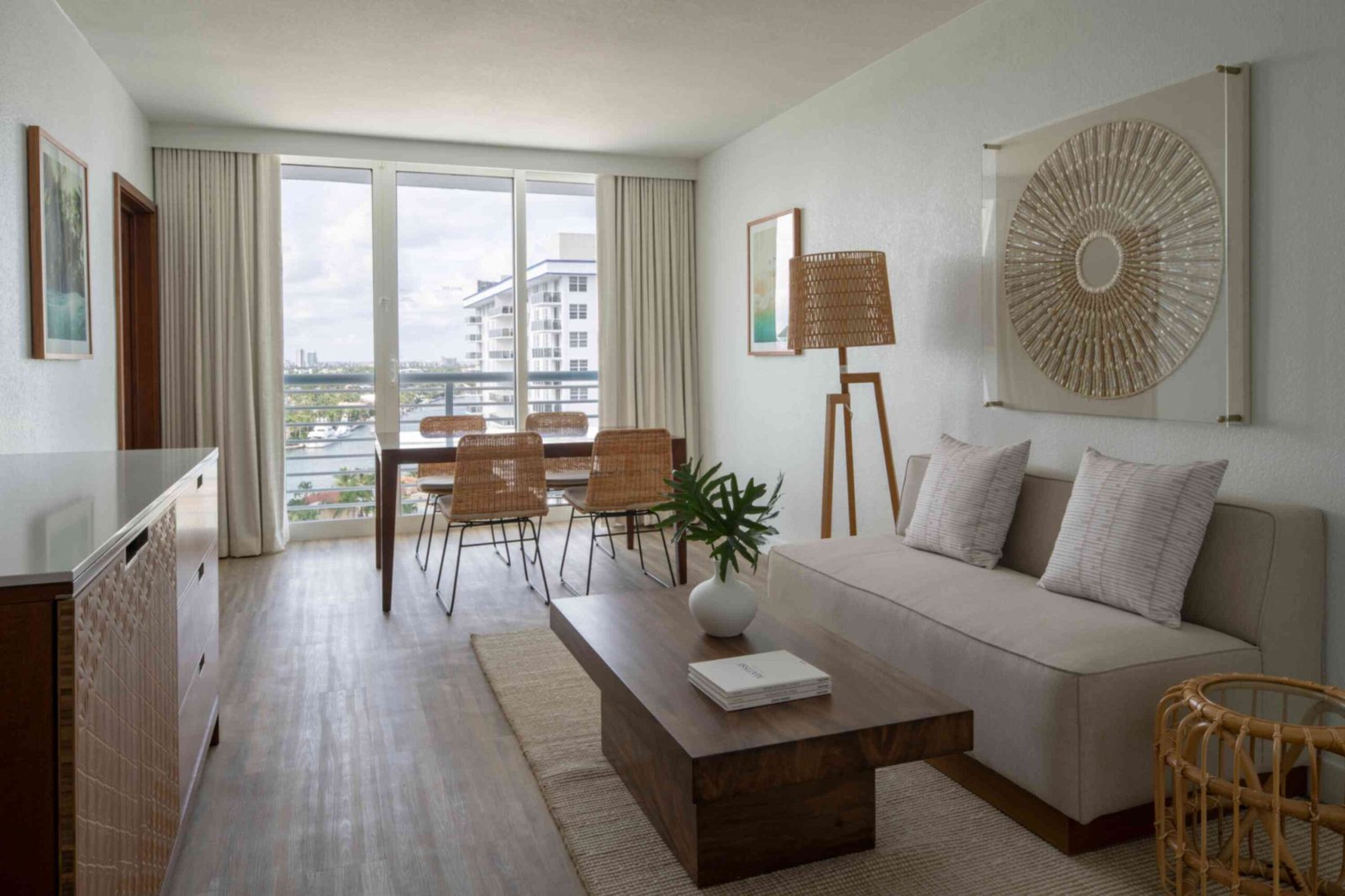 A bright hotel suite living area with a view of a city waterway through large glass sliding doors. The room features a light grey sofa with textured throw pillows, a solid wood coffee table, a woven floor lamp, and a large circular string art piece hanging on the wall. In the background, a wooden dining table is paired with four woven chairs next to the balcony.