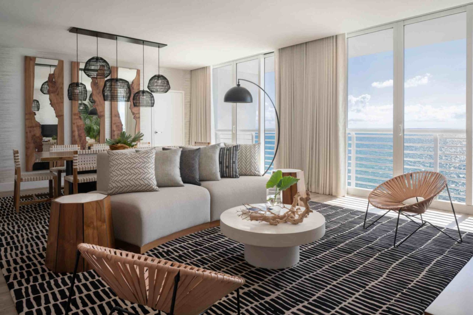 A sunlit hotel suite living room with panoramic ocean views through massive floor-to-ceiling glass doors. A large grey sectional sofa with textured throw pillows sits on a black and white patterned area rug alongside a round white coffee table and two woven accent chairs. In the background, a wooden dining table sits beneath black mesh pendant lights next to live-edge wood wall mirrors.