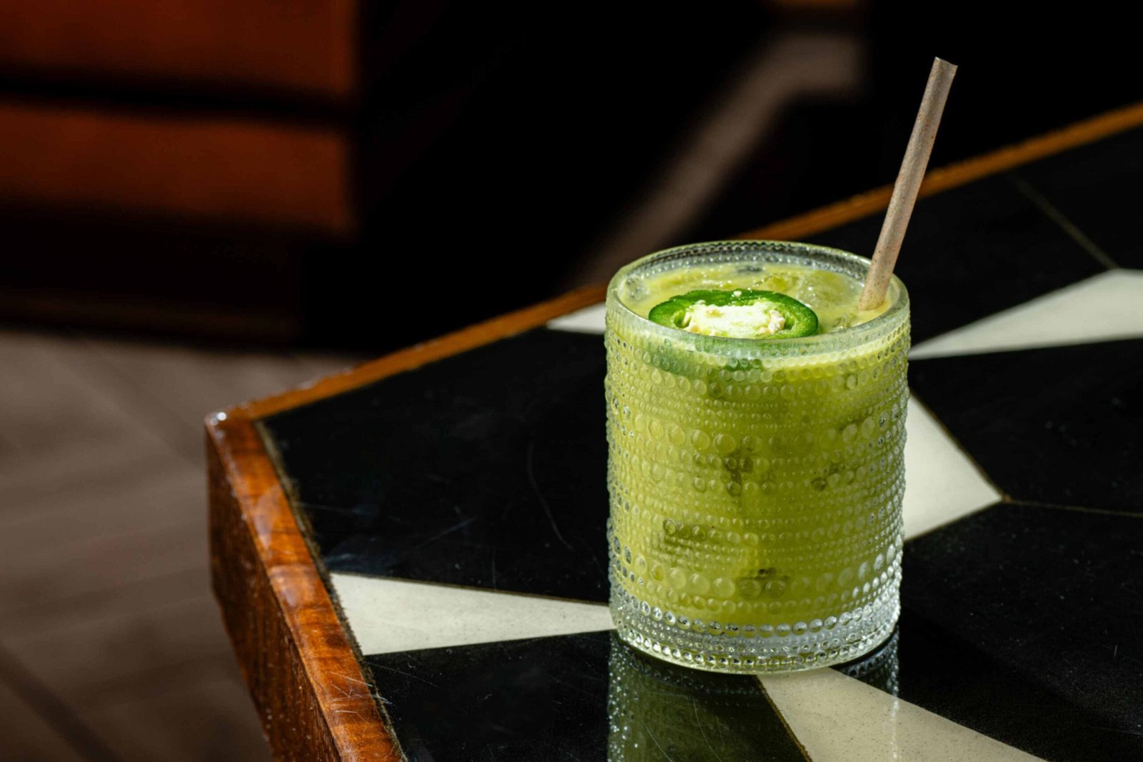 A green mezcal cocktail served in a textured, dotted glass on a black and white geometric patterned tabletop. The drink includes a paper straw and a fresh green jalapeño slice floating on top as a garnish.