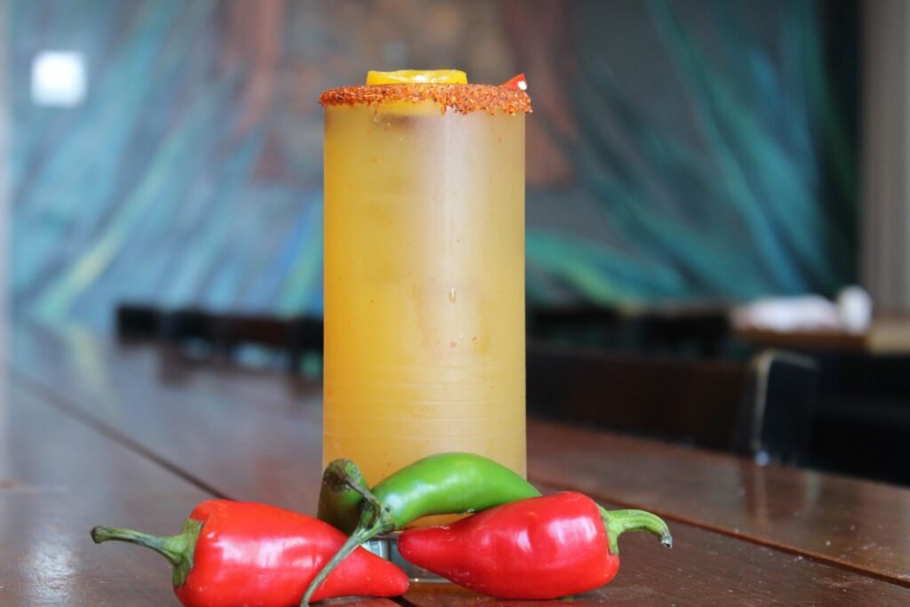 A tall iced cocktail in a frosted glass with a chili powder rim sits on a dark wood table. A yellow citrus slice floats at the top, and three whole chili peppers two red and one green rest at the base of the glass against a blurred blue background.