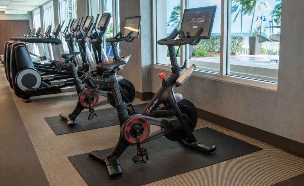 A row of modern Peloton stationary bikes inside a hotel gym, positioned on black mats next to a large window. In the background, other cardio machines line the room, while the window looks out toward an outdoor patio with palm trees and the ocean.