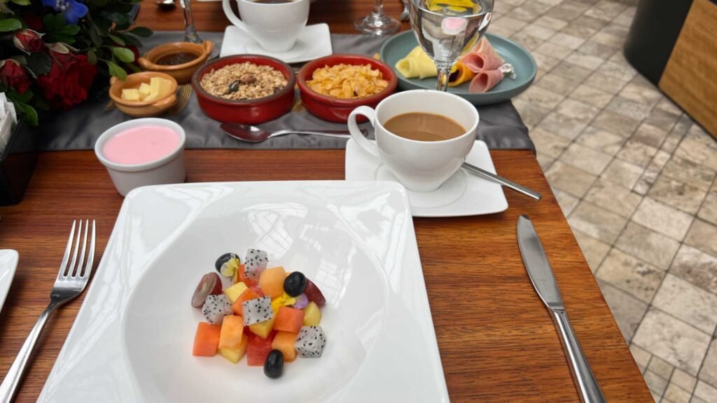 Close up overhead view of a gourmet breakfast spread on a wooden table featuring a central plate of diced fresh tropical fruit alongside coffee cups yogurt granola and a plate of ham and cheese