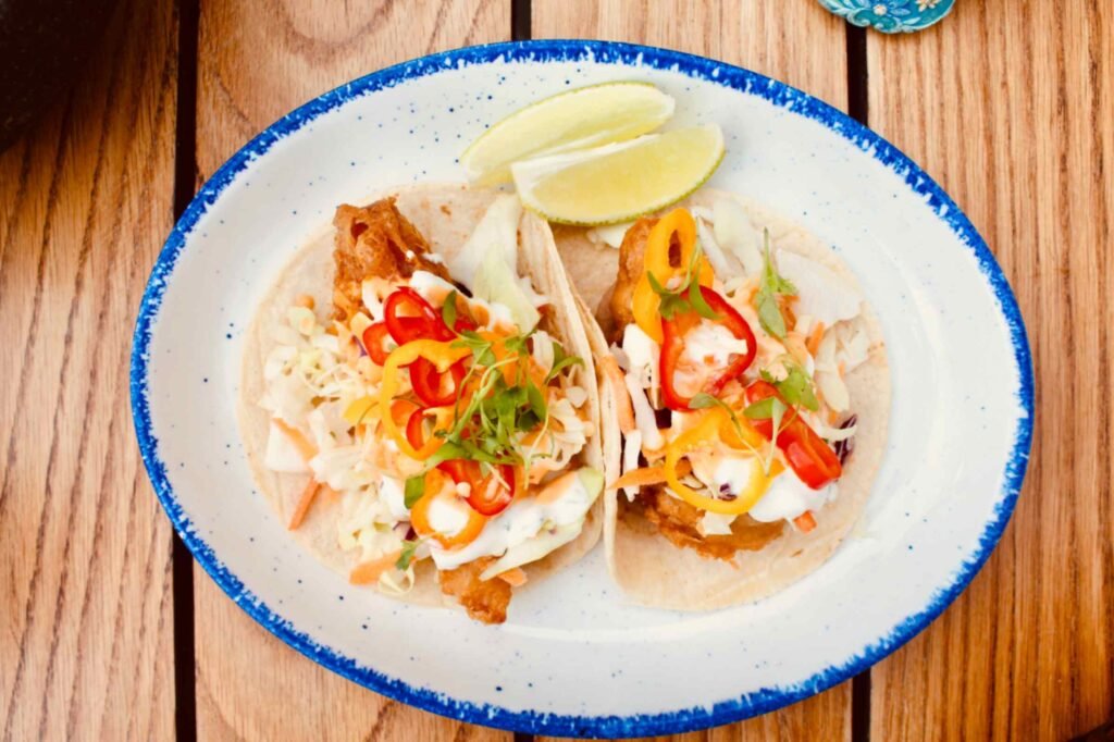 A top down view of two fish tacos served on soft corn tortillas, placed on a white plate with a speckled blue rim. The tacos are loaded with crispy battered fish, shredded cabbage slaw, crema, microgreens, and sliced red and yellow peppers, accompanied by two fresh lime wedges on a light wood tabletop.
