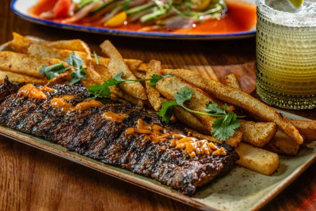 A grilled skirt steak dish served on an elongated ceramic platter alongside seasoned thick-cut french fries. The steak is drizzled with a savory orange sauce and garnished with cilantro sprigs, set on a wooden table with a salted-rim cocktail and a seafood dish in the background.
