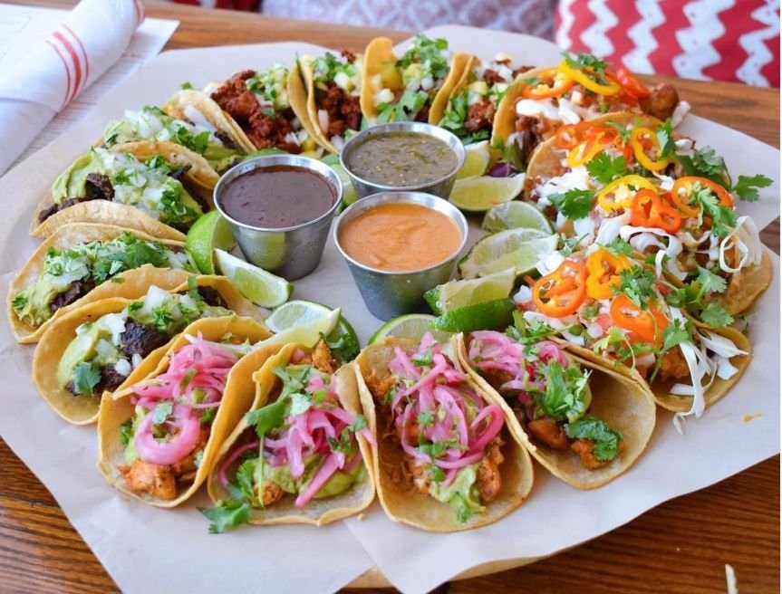 A large circular platter filled with an assortment of street tacos arranged in a ring. The platter includes tacos with grilled meat, pickled pink onions, fresh cabbage slaw, chopped peppers, cilantro, and guacamole, surrounding three metal cups filled with different salsas and fresh lime wedges on a wooden table.