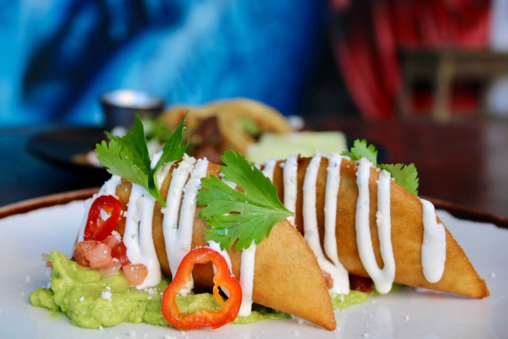 A close-up view of fried empanadas served on a bed of fresh guacamole and pico de gallo. The golden-brown empanadas are drizzled with white crema, sprinkled with crumbled cheese, and garnished with fresh cilantro leaves and sliced red peppers.