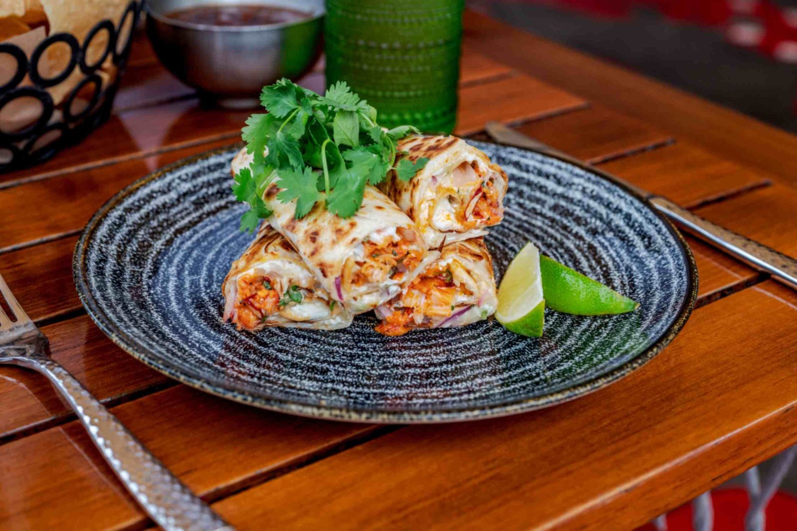 A plate of lobster quesadillas cut into rolls and stacked on a textured black and white ceramic dish. The quesadillas are filled with shredded lobster meat, melted cheese, and red onions, topped with a bunch of fresh cilantro and served with a lime wedge on a wooden table.