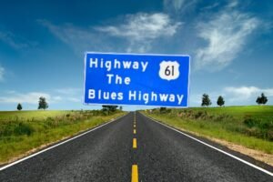 An image of a large, rectangular blue roadside sign with white text that reads "Highway 61 / The / Blues Highway". The number "61" is enclosed inside a white shield icon, representing a U.S. Route shield. The sign is supported by three metal posts and stands along the side of a paved highway. In the background, there is a grassy slope, a line of green trees, and a clear, light blue sky.