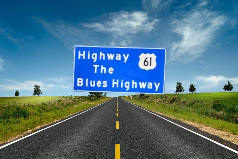 An image of a large, rectangular blue roadside sign with white text that reads "Highway 61 / The / Blues Highway". The number "61" is enclosed inside a white shield icon, representing a U.S. Route shield. The sign is supported by three metal posts and stands along the side of a paved highway. In the background, there is a grassy slope, a line of green trees, and a clear, light blue sky.
