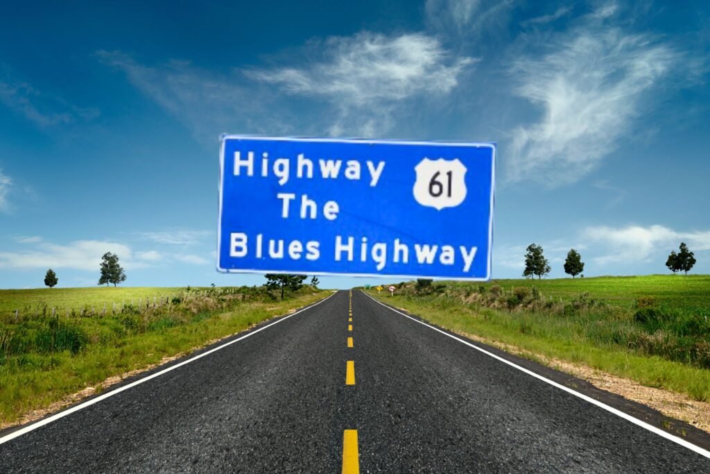 An image of a large, rectangular blue roadside sign with white text that reads "Highway 61 / The / Blues Highway". The number "61" is enclosed inside a white shield icon, representing a U.S. Route shield. The sign is supported by three metal posts and stands along the side of a paved highway. In the background, there is a grassy slope, a line of green trees, and a clear, light blue sky.