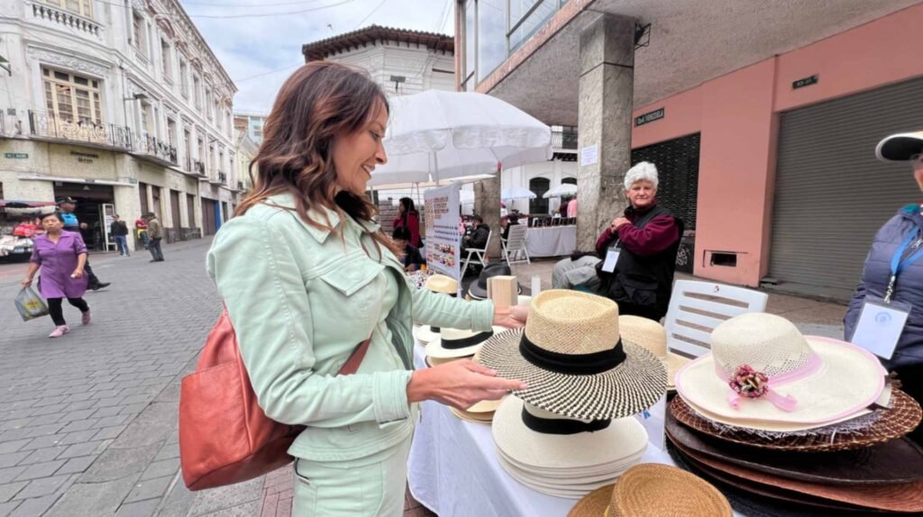 A woman with long wavy brown hair wearing a light green jacket smiles as she browses hand woven straw hats at a sidewalk market stall on a historic cobblestone street in Quito Ecuador