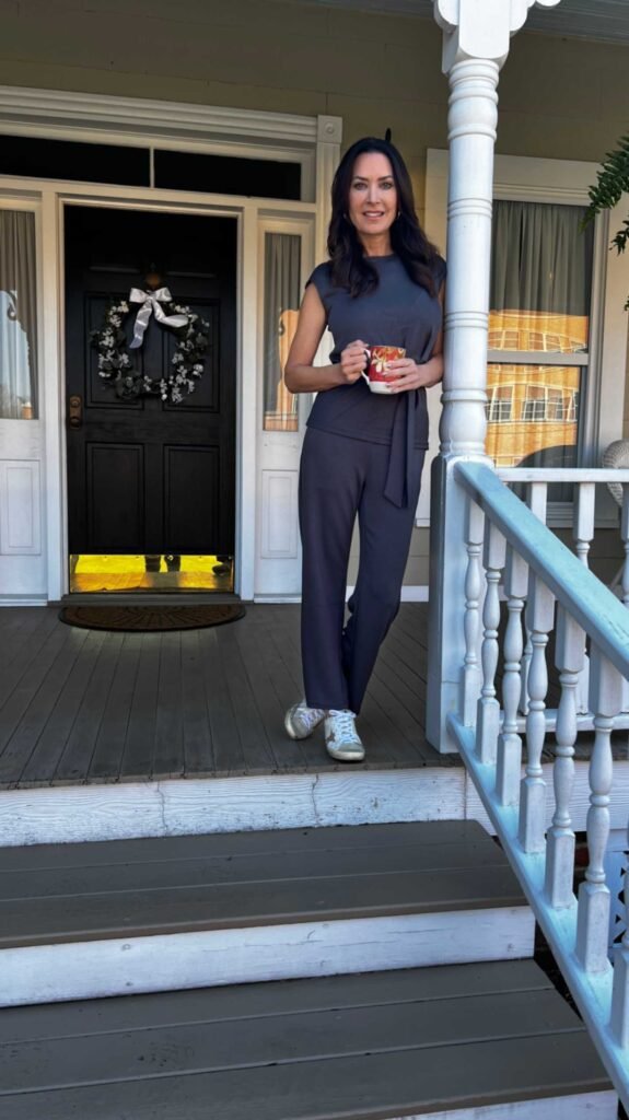 A woman with dark hair leans against a white porch pillar outside a house with a black front door. She is smiling and holding a red patterned coffee mug. She is wearing a dark gray or charcoal sleeveless top with a waist tie, matching dark gray pants, and white sneakers.