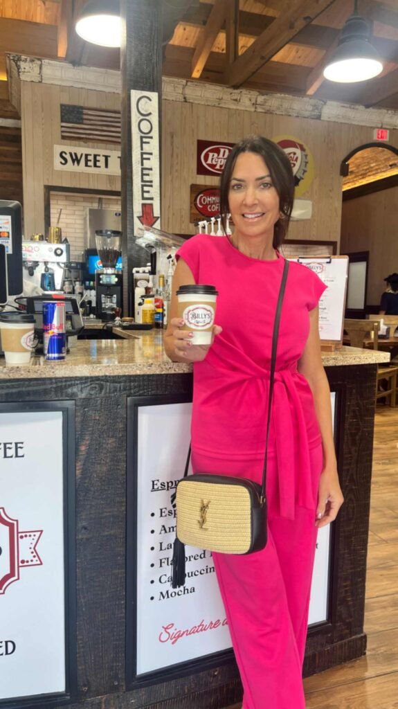 A woman with dark hair stands inside a coffee shop smiling, holding a white paper coffee cup. She is wearing a bright fuchsia pink short-sleeve top with a front tie at the waist and matching pink pants. A small woven straw crossbody bag with a black leather trim and gold logo hangs across her shoulder. A wrinkle-free Amazon sets that keep me camera-ready all summer.