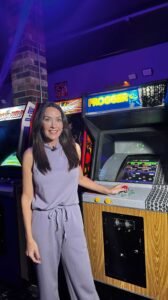 A woman with dark hair stands in an arcade, smiling with one hand resting on the controls of a vintage Frogger arcade game. She is wearing a sleeveless lavender purple top and matching drawstring jogger-style pants.