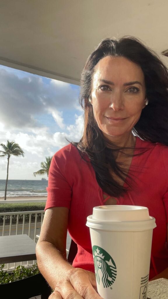 A woman with long dark hair and a red short sleeved shirt sitting at an outdoor patio table with a white Starbucks coffee cup in the foreground. In the background, a view reveals palm trees, a sandy beach, and ocean waves rolling in under a partly cloudy sky.