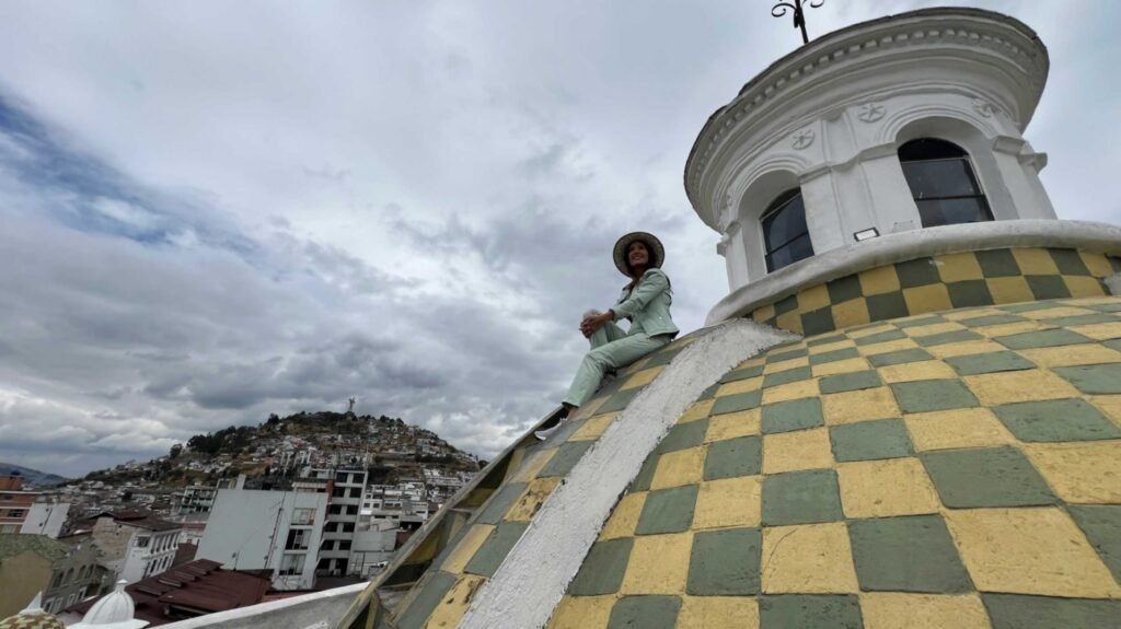 Low angle view of a woman in a light green outfit and sun hat sitting on the slope of a large yellow and green checkered dome belonging to the Quito Metropolitan Cathedral under a dramatic cloudy sky