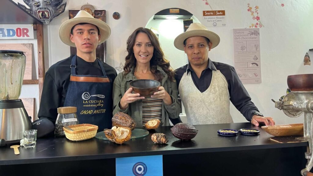 A woman standing behind a black counter between two men wearing traditional straw hats during a chocolate workshop featuring raw cacao pods and a blender in Quito Ecuador