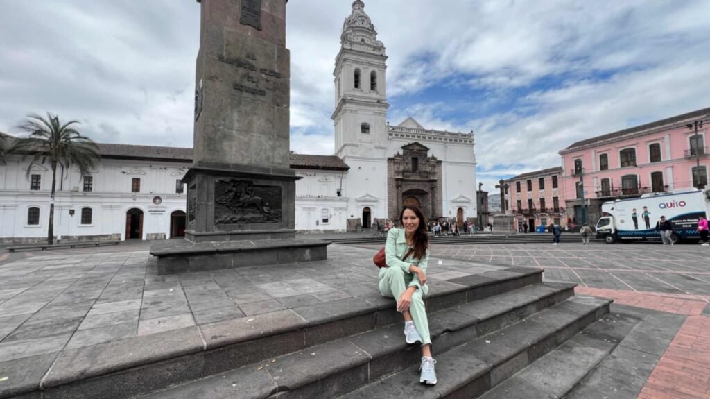 A woman in a light green outfit sitting on the stone steps of Plaza de Santo Domingo in Quito Ecuador with a massive stone monument on the left and a white colonial church tower in the background